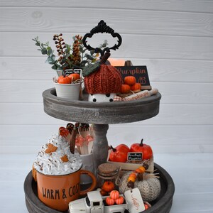 May include: A two-tiered wooden tray with fall-themed decorations. The top tier features a knitted pumpkin, mini pumpkins, and greenery. The bottom tier includes a mug with whipped cream, a toy truck with pumpkins, and a sign that says "Warm & Cozy."