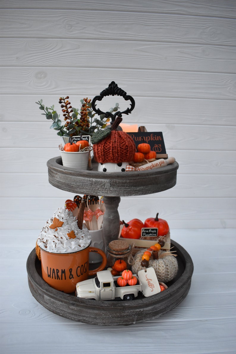 May include: A two-tiered wooden tray with fall-themed decorations. The top tier features a knitted pumpkin, mini pumpkins, and greenery. The bottom tier includes a mug with whipped cream, a toy truck with pumpkins, and a sign that says "Warm & Cozy."