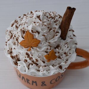 May include: A ceramic mug filled with a whipped cream topping, cocoa powder, and a cinnamon stick. The mug is light orange with the words "Warm & Cozy" on the side, and two orange leaf decorations.