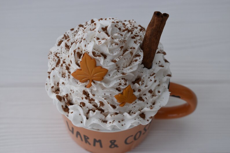 May include: A ceramic mug filled with a whipped cream topping, cocoa powder, and a cinnamon stick. The mug is light orange with the words "Warm & Cozy" on the side, and two orange leaf decorations.