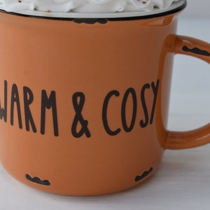 May include: An orange enamel mug with the words "WARM & COSY" in black lettering. The mug has a black rim and handle, and is filled with a white substance, possibly whipped cream. The mug has a distressed look.