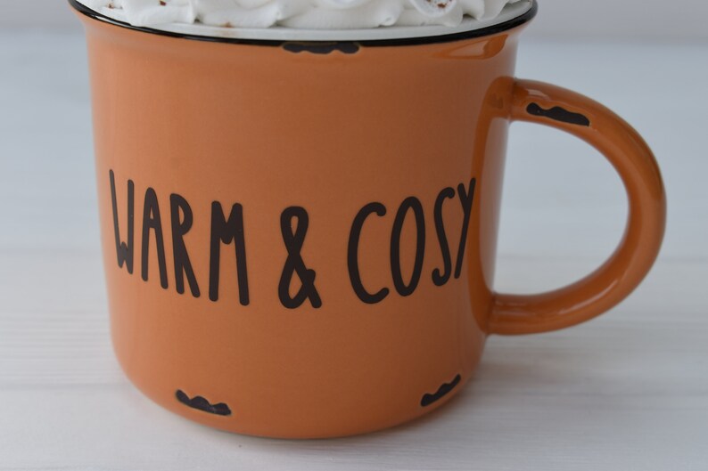 May include: An orange enamel mug with the words "WARM & COSY" in black lettering. The mug has a black rim and handle, and is filled with a white substance, possibly whipped cream. The mug has a distressed look.
