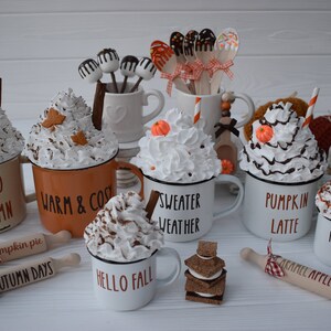 May include: A collection of fall-themed decorative mugs with whipped cream toppings, cinnamon sticks, and leaf accents. Some mugs have text like "Hello Autumn" and "Pumpkin Spice." Rolling pins with text like "Caramel Apples" are also present.