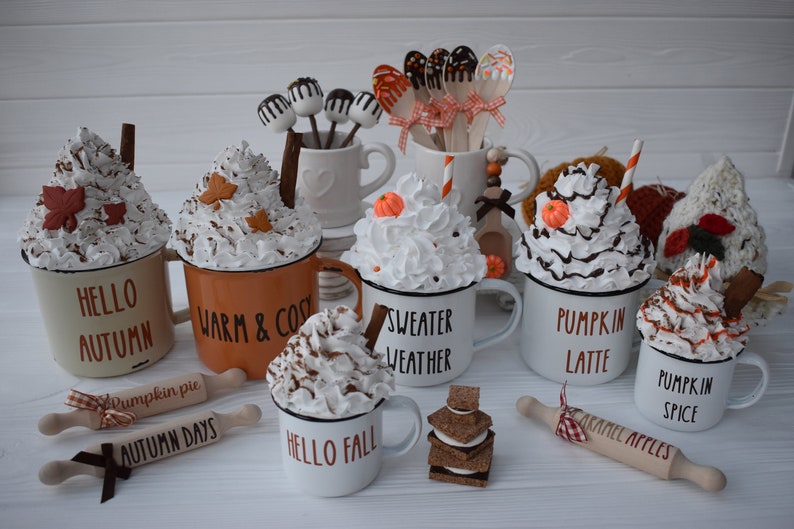 May include: A collection of fall-themed decorative mugs with whipped cream toppings, cinnamon sticks, and leaf accents. Some mugs have text like "Hello Autumn" and "Pumpkin Spice." Rolling pins with text like "Caramel Apples" are also present.