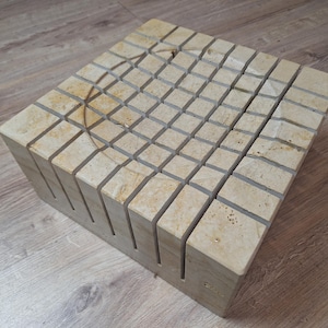 May include: A square, beige-colored, decorative tile block with a grid pattern and a circular indentation in the center.