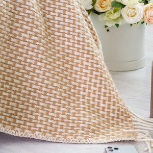 May include: A beige and white woven throw blanket with fringe edges. The blanket is draped over a white surface and a white vase with flowers is visible in the background.