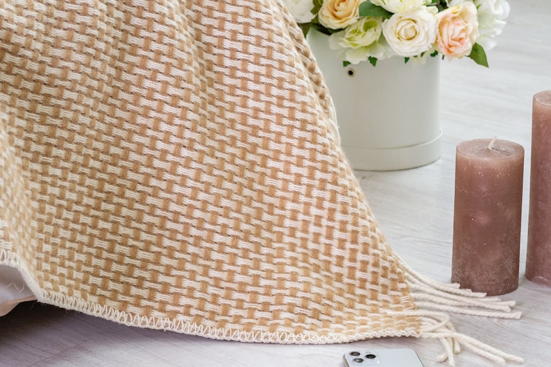 May include: A beige and white woven throw blanket with fringe edges. The blanket is draped over a white surface and a white vase with flowers is visible in the background.