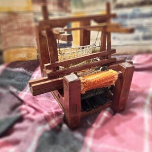 May include: A wooden loom with a woven orange and white fabric on the warp. The loom is sitting on a red and black plaid blanket.