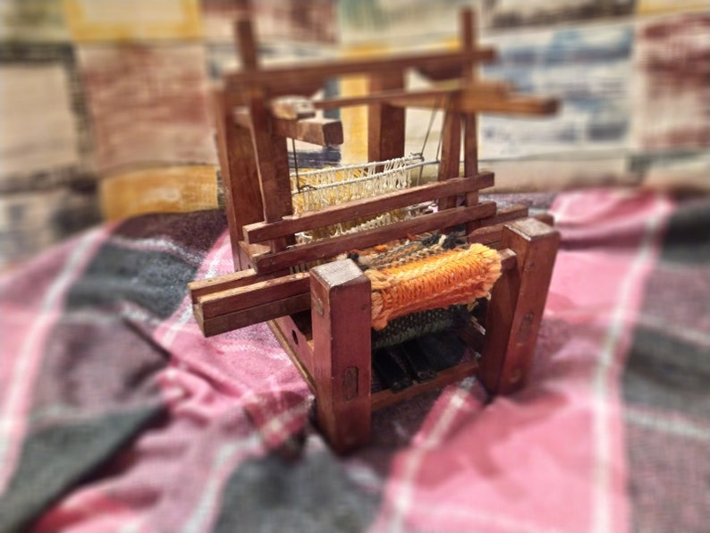 May include: A wooden loom with a woven orange and white fabric on the warp. The loom is sitting on a red and black plaid blanket.