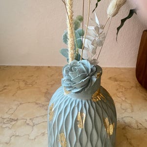 Peut inclure: Un vase bleu clair avec un motif ondulé texturé et des accents de feuilles d'or. Le vase contient des fleurs séchées et de la verdure, notamment des épis de blé et des succulentes. Le vase a un dessus en forme de rose.