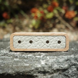 May include: A rectangular wooden box with a light gray leather insert. The insert has four evenly spaced holes. The wood has a light, natural finish. The box is sitting on a gray stone surface.