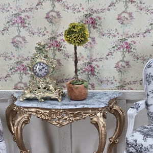 May include: A miniature antique-style scene featuring a gold-colored ornate clock and a small topiary tree in a terracotta pot. The clock has a white face with black Roman numerals. The table has a marble top and gold legs. The background is floral wallpaper.