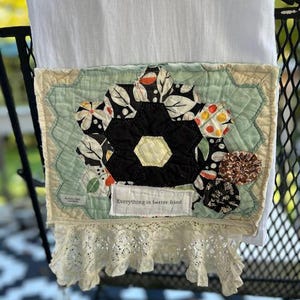 May include: A white kitchen towel featuring a quilted patch with a black, white, and orange floral hexagon pattern. A small white fabric label reads "Everything is better fried." The bottom edge is finished with a cream-colored lace trim.