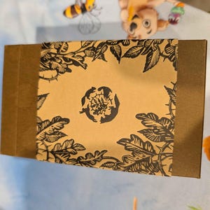 May include: A brown rectangular box with a decorative band featuring a floral design in black. The band is centered with a black floral emblem. The box is on a light blue surface with cartoon characters.