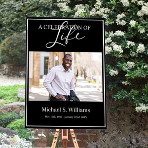 Op de afbeelding: Een zwart en zilveren ingelijst bord met de tekst "A Celebration of Life" in zilveren schrift. Het bord bevat ook een foto van een lachende man, de tekst "Michael S. Williams" en de data "May 15th, 1985 - January 22nd, 20XX".