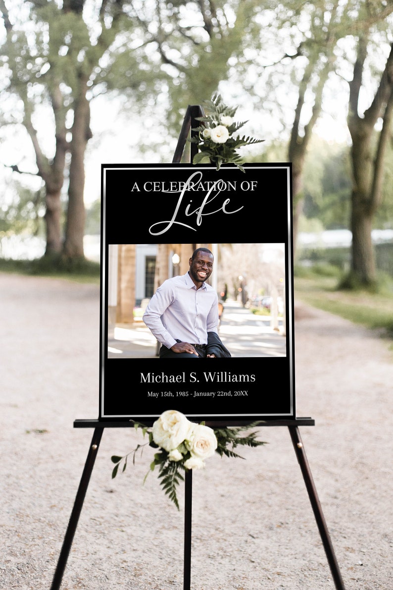 Op de afbeelding: Een zwart-wit bord met een zilveren inscriptie die "A Celebration of Life" luidt, met een foto van een man en de tekst "Michael S. Williams, May 15th, 1985 - January 22nd, 20XX".