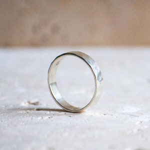 Könnte beinhalten: Ein silberner Ring mit gehämmertem Muster. Der Ring ist rund und hat ein flaches Band. Der Ring liegt auf einer weißen Oberfläche. Der Hintergrund ist in einem neutralen Beigeton gehalten.