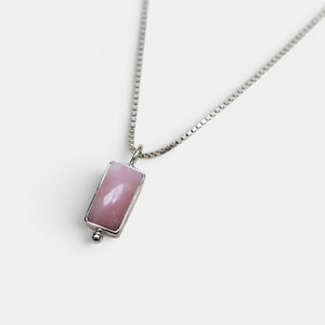 Peut inclure: Un collier à chaîne en argent avec un pendentif rectangulaire orné d'une pierre rose clair lisse sertie dans un cadre en argent. Le pendentif est fixé à la chaîne par une petite bélière en argent, offrant un design sobre et épuré.