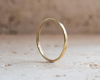 May include: A delicate, thin gold ring stands upright against a neutral background. The ring's simple design and polished finish reflect the light, highlighting its circular shape. This minimalist piece is suitable for everyday wear.