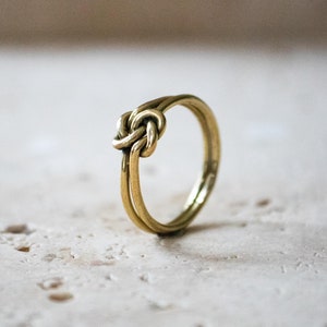 Può includere: Un anello d'oro con un designo a nodo. L'anello è composto da due bande di metallo intrecciate per creare un nodo.