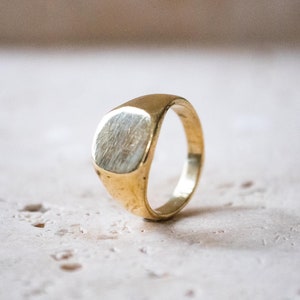 Könnte beinhalten: Ein goldener Siegelring mit einer glatten, abgerundeten Oberseite. Der Ring ist aus Metall und hat ein schlichtes, klassisches Design.