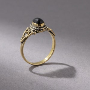 Könnte beinhalten: Ein goldener Ring mit einem schwarzen Edelstein, der in einer Zargenfassung gefasst ist. Der Ring hat ein zartes, verziertes Design mit einem gedrehten Band und einem dekorativen Motiv auf der Schiene.