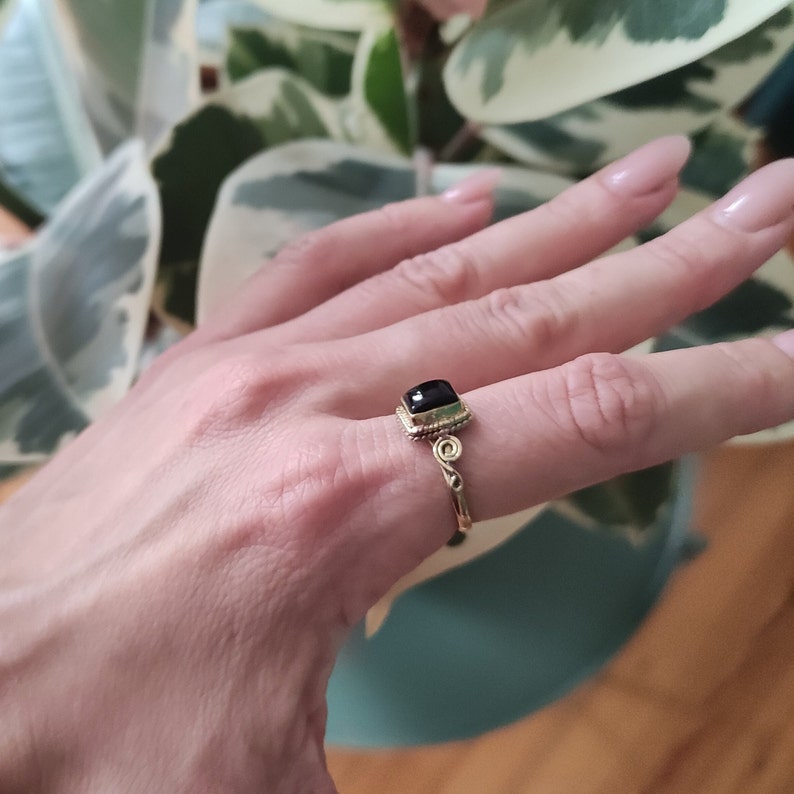 K&ouml;nnte beinhalten: Ein goldener Ring mit einem quadratischen schwarzen Edelstein, der in einen dekorativen Rahmen gefasst ist. Der Ring hat ein verdrehtes Band.