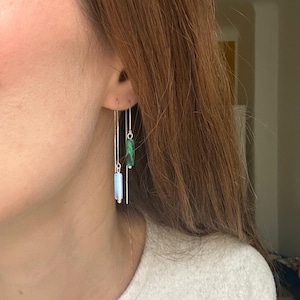 Pode incluir: Um par de brincos pendurados com um design minimalista. Cada brinco apresenta uma delicada corrente de prata que suspende uma pedra retangular. Uma pedra é azul clara e a outra verde. Os brincos são mostrados em um fundo neutro.