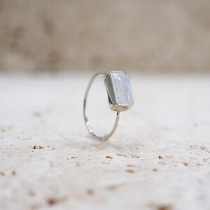 Puede incluir: Un anillo de plata con una piedra preciosa rectangular y clara. El anillo tiene una banda delgada y delicada, y la piedra est&aacute; engastada en un estilo simple y minimalista. La piedra tiene una calidad sutil e iridiscente.