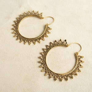 Könnte beinhalten: Ein Paar goldfarbene Creolen. Jeder Ohrring hat ein kreisförmiges Design mit aufwendigen Filigranarbeiten und einem sonnenstrahlenartigen Muster am äußeren Rand. Die Ohrringe haben einen Hakenverschluss.