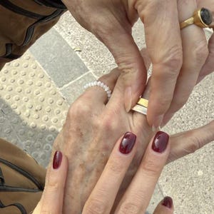 May include: Close-up of two hands with a gold ring on one finger and a pearl bracelet on the other. The person on the left is placing a gold band on the ring finger of the person on the right.