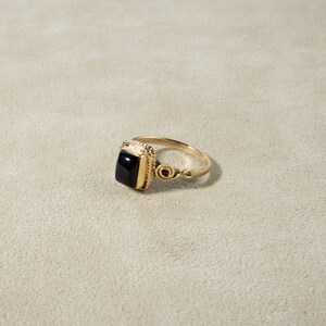 K&ouml;nnte beinhalten: Ein goldener Ring mit einem schwarzen quadratischen Edelstein, der in einem dekorativen Rahmen gefasst ist. Der Ring hat ein zartes, wirbelndes Design an der Band.