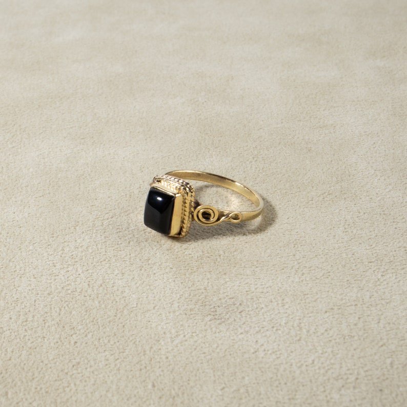 K&ouml;nnte beinhalten: Ein goldener Ring mit einem schwarzen quadratischen Edelstein, der in einem dekorativen Rahmen gefasst ist. Der Ring hat ein zartes, wirbelndes Design an der Band.