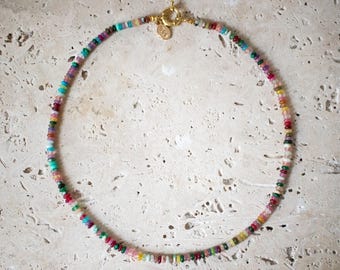Può includere: Una collana di perline multicolore con chiusura dorata. La collana presenta piccole perline rotonde in tonalità di rosso, verde, blu, giallo e viola. La collana è esposta su una superficie chiara e strutturata.