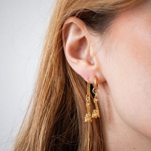 Può includere: Orecchini color oro dal design unico. Uno è un piccolo cerchio, l'altro ha una delicata catena con un elemento decorativo. Gli orecchini sono indossati sull'orecchio di una persona, con i capelli visibili.