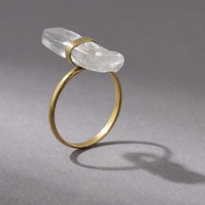 Op de afbeelding: Een gouden ring met een helder kwartskristal erop. Het kristal heeft een ruwe, natuurlijke vorm.