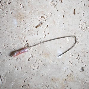 Könnte beinhalten: Eine zarte silberne Kette mit einem Anhänger aus rosa und weißer Perle. Die Kette hängt auf einer hellbeigen Oberfläche.