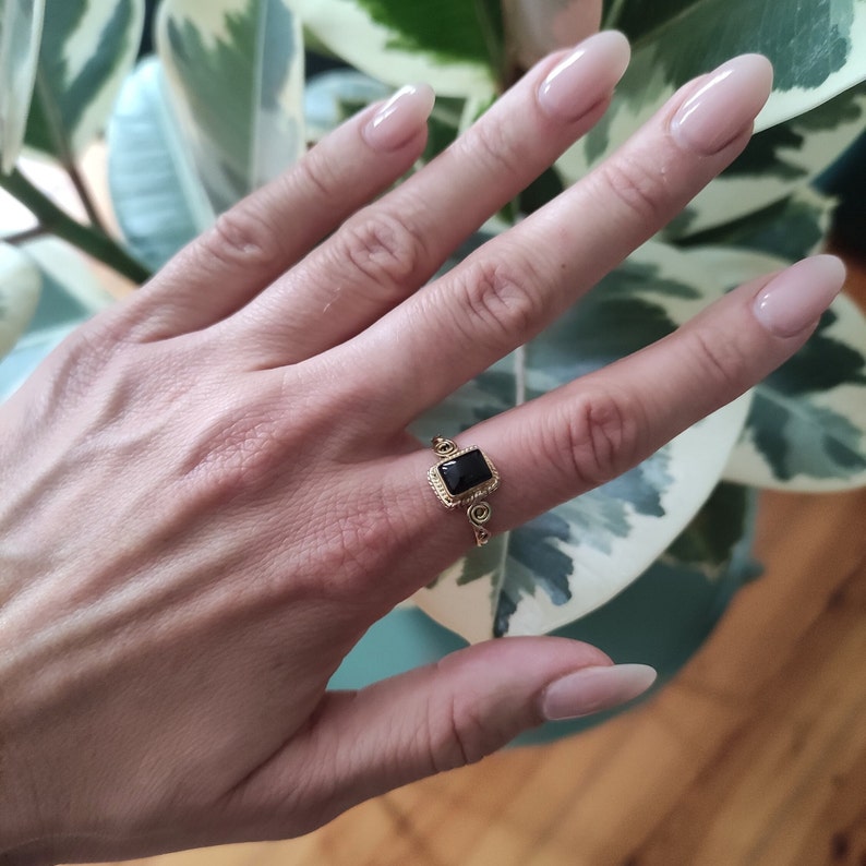 K&ouml;nnte beinhalten: Ein goldener Ring mit einem schwarzen quadratischen Edelstein, der in einem dekorativen Rahmen gefasst ist. Der Ring wird an einem Finger getragen.