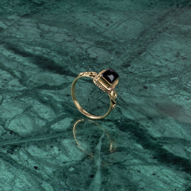 K&ouml;nnte beinhalten: Ein goldener Ring mit einem schwarzen quadratischen Edelstein, der in eine dekorative goldene Fassung gefasst ist.