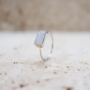 Puede incluir: Un anillo de plata con una piedra preciosa rectangular e iridiscente. La piedra tiene un suave tono blanco y azul perlado. La banda del anillo es fina y delicada, resaltando el dise&ntilde;o de la gema. Una joya minimalista.