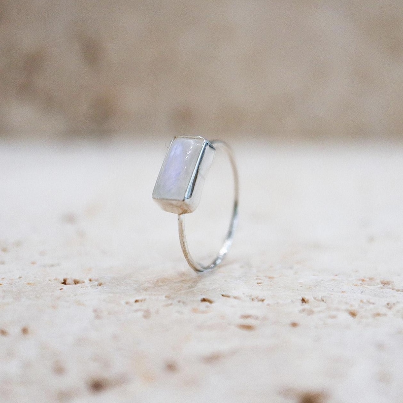 Puede incluir: Un anillo de plata con una piedra preciosa rectangular e iridiscente. La piedra tiene un suave tono blanco y azul perlado. La banda del anillo es fina y delicada, resaltando el dise&ntilde;o de la gema. Una joya minimalista.