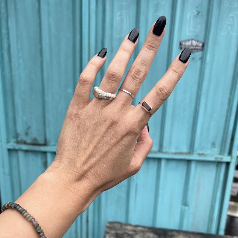May include: A hand wearing three silver rings. The ring on the index finger is a pearl ring. The ring on the middle finger is a thin band. The ring on the pinky finger has a rectangular brown stone. The nails are painted black.