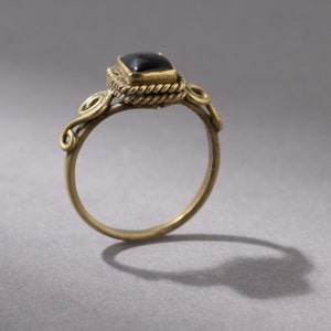 K&ouml;nnte beinhalten: Ein goldener Ring mit einem schwarzen Edelstein, der in einer quadratischen Fassung sitzt. Der Ring hat ein zartes, verziertes Design mit gedrehten Drahtdetails.