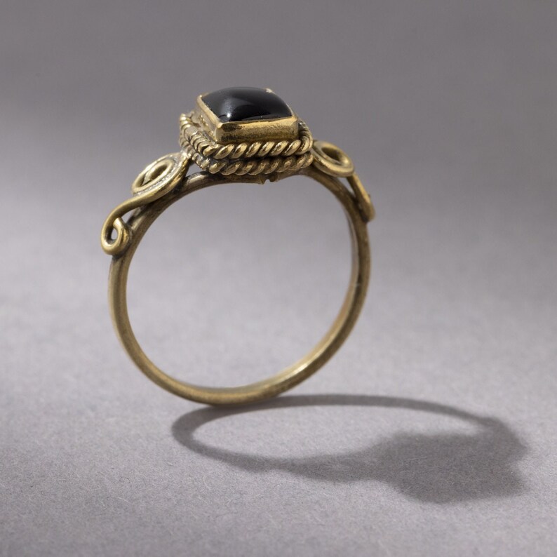 K&ouml;nnte beinhalten: Ein goldener Ring mit einem schwarzen Edelstein, der in einer quadratischen Fassung sitzt. Der Ring hat ein zartes, verziertes Design mit gedrehten Drahtdetails.