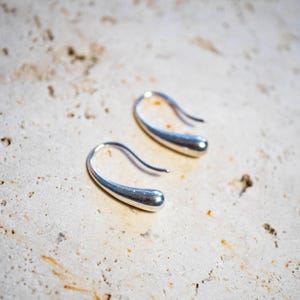 May include: A pair of silver teardrop earrings. The earrings feature a curved hook design and a smooth, polished finish. The teardrop shape is elongated and slightly rounded at the bottom. The earrings are displayed on a light-coloured, textured surface.