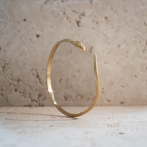 Puede incluir: Un brazalete de serpiente dorado. El brazalete es abierto, con la cabeza de la serpiente en un extremo y la cola en el otro. La cabeza y la cola de la serpiente tienen texturas detalladas, sobre un fondo neutro.