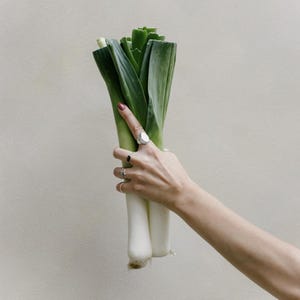K&ouml;nnte beinhalten: Eine Hand h&auml;lt einen Bund frischer gr&uuml;ner Lauchzwiebeln mit wei&szlig;en Stielen. Die Hand tr&auml;gt mehrere Ringe.