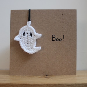May include: A handmade Halloween card featuring a crocheted white ghost charm hanging from a black ribbon. The card is a square shape with a brown paper texture, and the word "Boo!" is written in black.