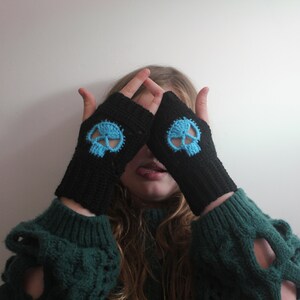 May include: Black fingerless gloves with a light blue skull design. The gloves are made of a knitted material. The skull design is centered on the back of the hand. The gloves are worn over a green sweater.