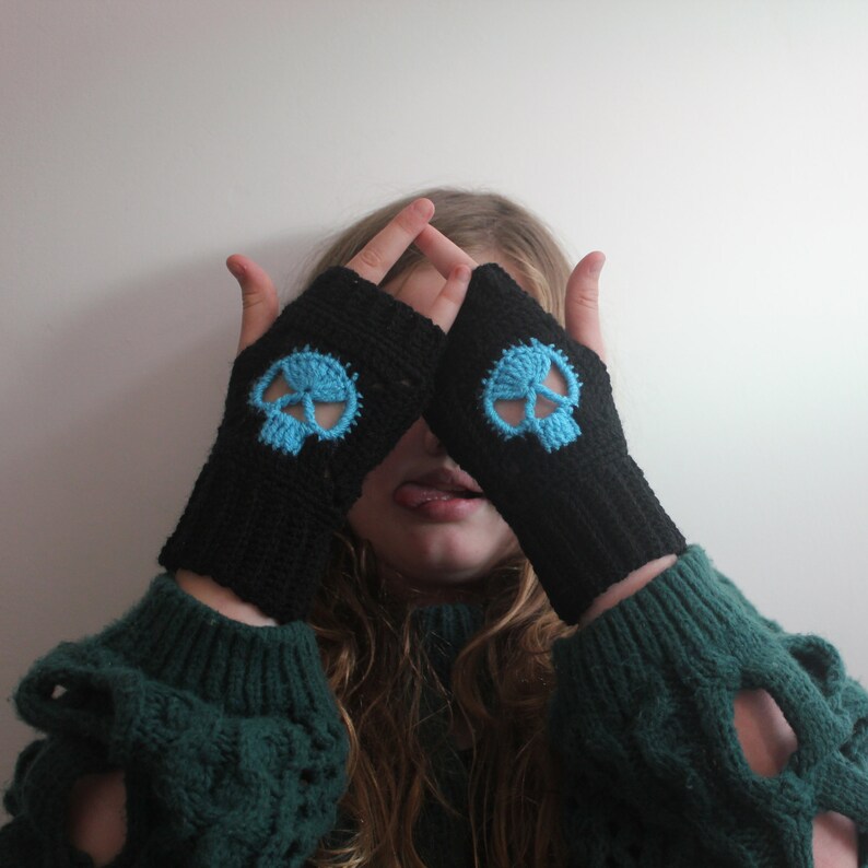 May include: Black fingerless gloves with a light blue skull design. The gloves are made of a knitted material. The skull design is centered on the back of the hand. The gloves are worn over a green sweater.